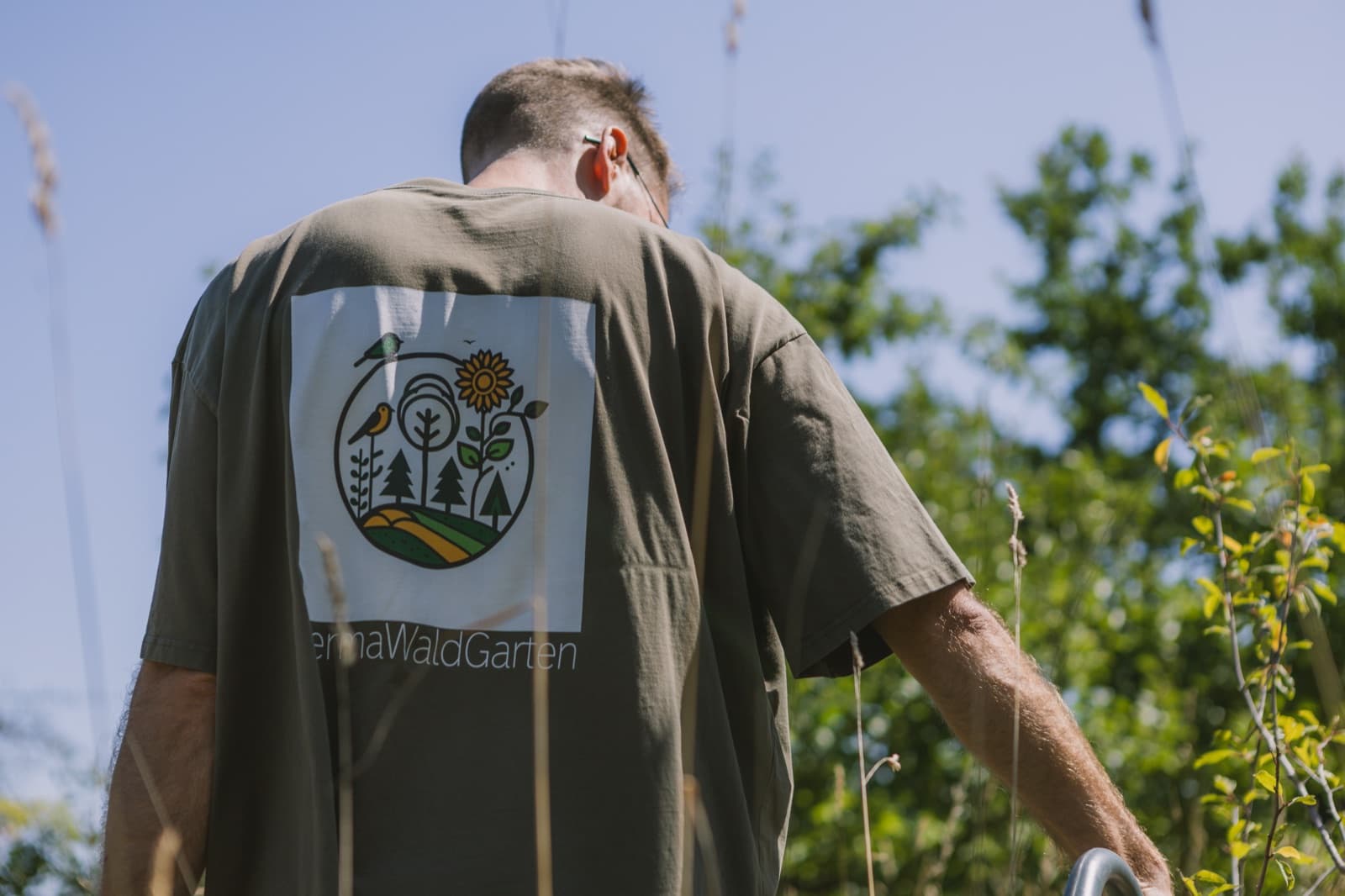 PermaWaldGarten-Logo mit Vogel, Sonnenblume und Bäumen auf einem T-Shirt